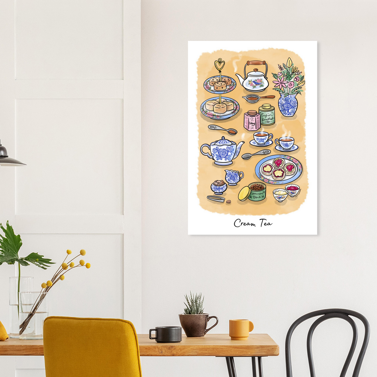 Wall art print titled 'Cream Tea' hangs on a white wall in a styled dining or living room setting. The artwork shows an illustrated tea party spread with teapots, cups, plates, flowers, and jars on a warm cream background. Great for blue and white home decor.