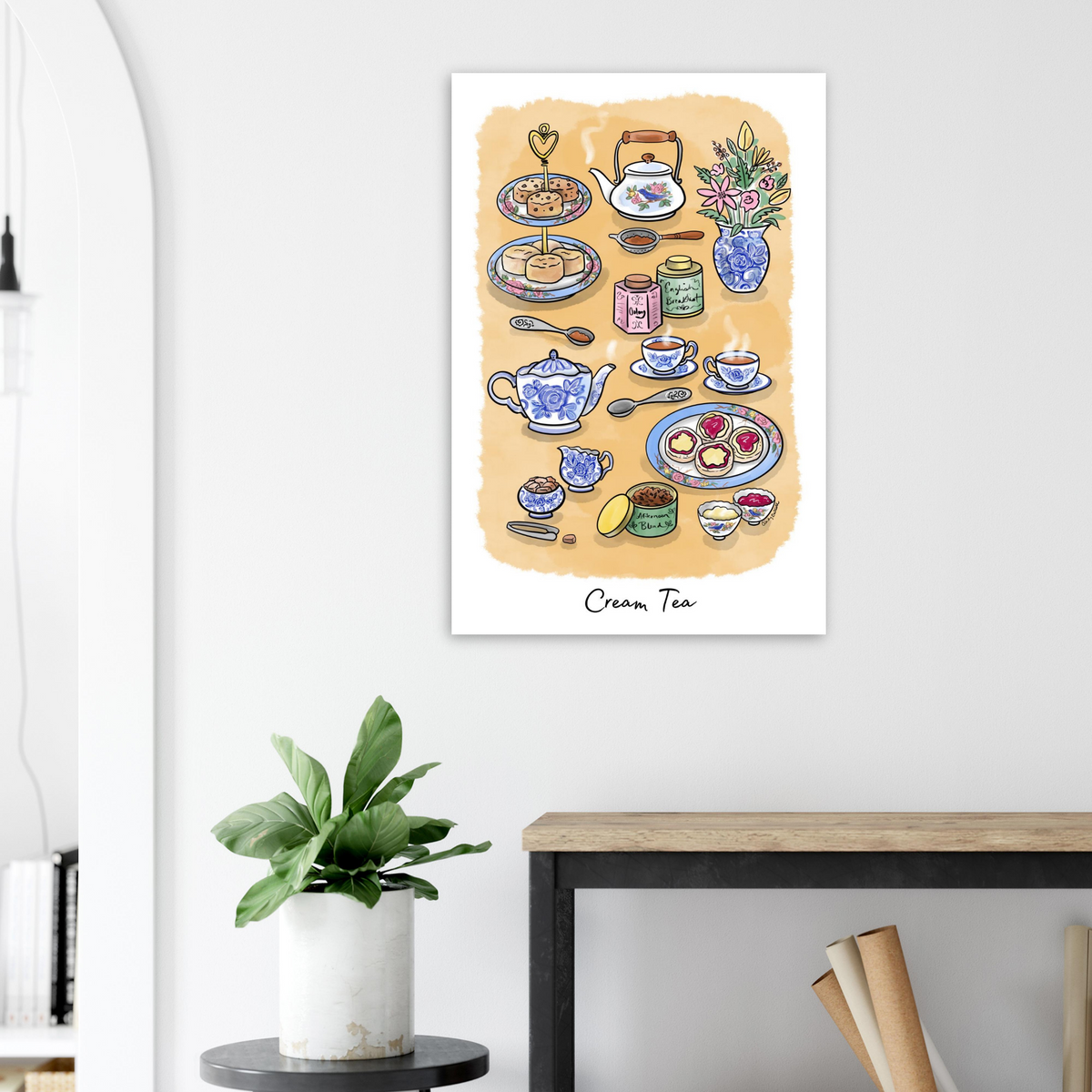 Art print featuring a colorful hand-drawn illustration of a cream tea tea party scene, displayed on a light gray wall above a wooden table with a potted plant.