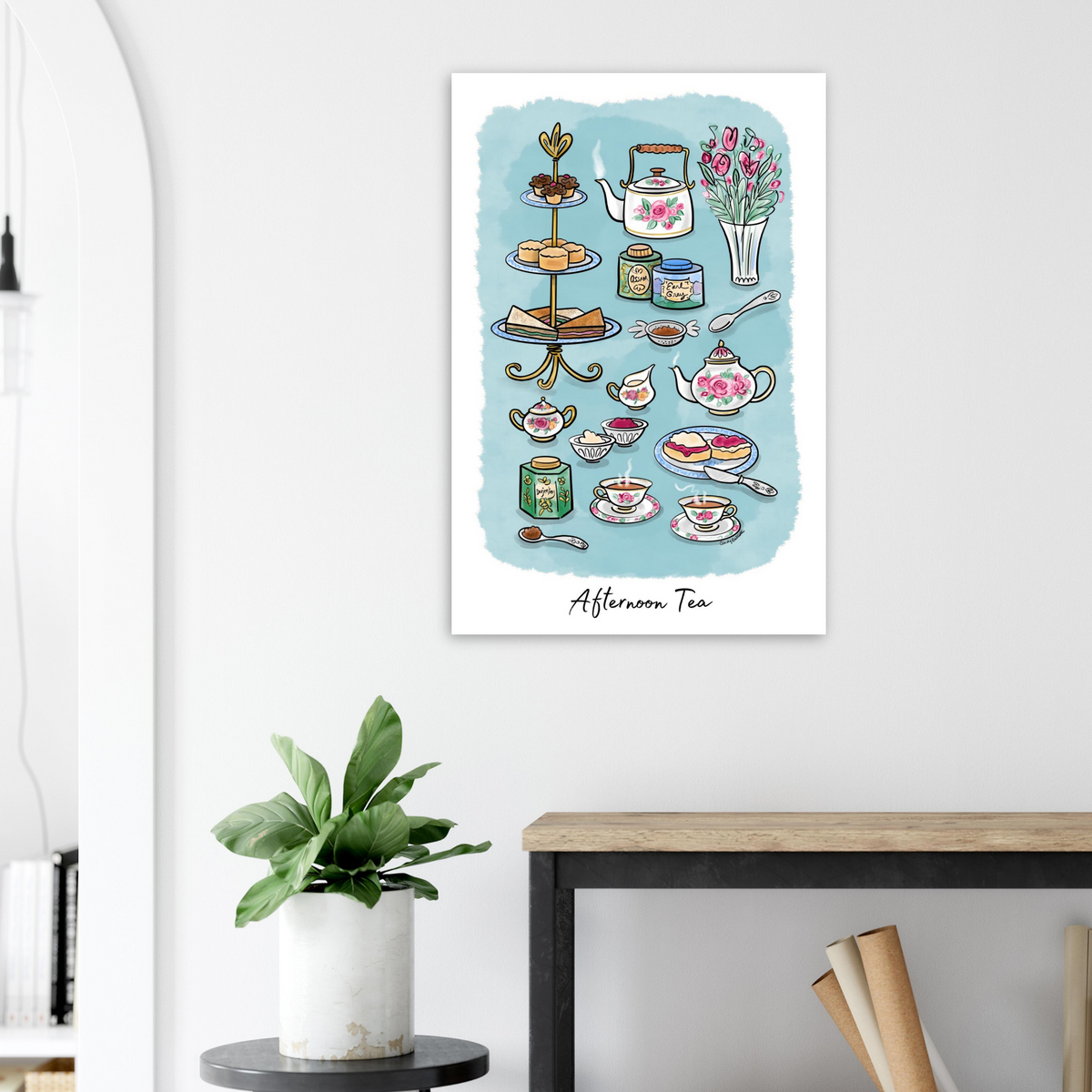 A vertical wall art print featuring a watercolor-style afternoon tea illustration, displayed on a white wall above a wooden table with a potted plant in a modern interior setting.